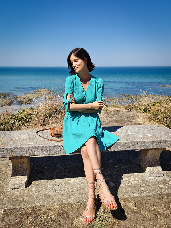 Une jeune femme brune est assise sur un banc devant la mer. Elle porte une robe d'une couleur turquoise très vive, qui rappelle celle du ciel et de la mer. La robe est en double gaze, elle semble légère et aérée.