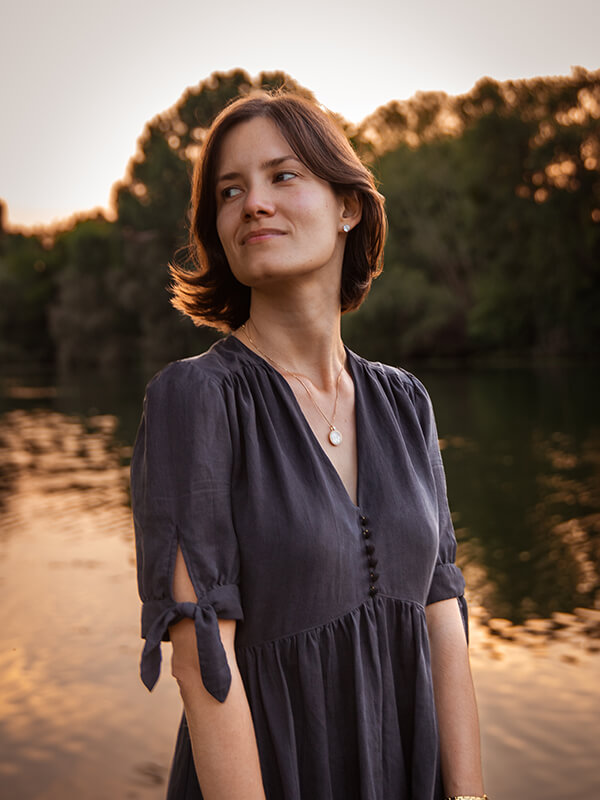 Une jeune femme brune porte une robe noire, délicatement froncée aux épaules, ornée de boutons recouverts du même tissu. Les manches sont courtes et nouées. En arrière-plan, le soleil se couche sur une rivière, baignant l'ensemble d'une lumière dorée.