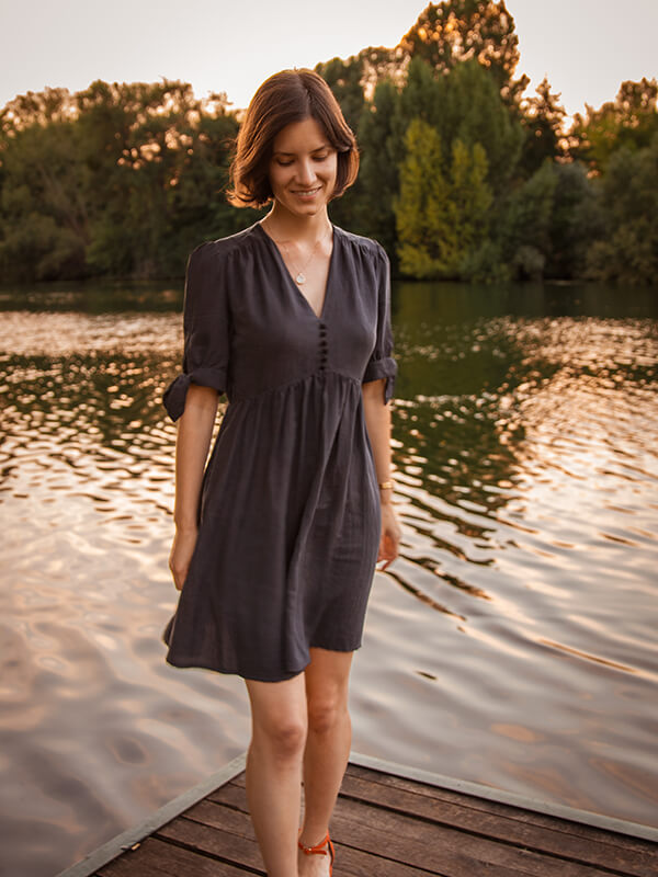 Une jeune femme brune porte une robe noire, délicatement froncée aux épaules, ornée de boutons recouverts du même tissu. Les manches sont courtes et nouées. En arrière-plan, le soleil se couche sur une rivière, baignant l'ensemble d'une lumière dorée.