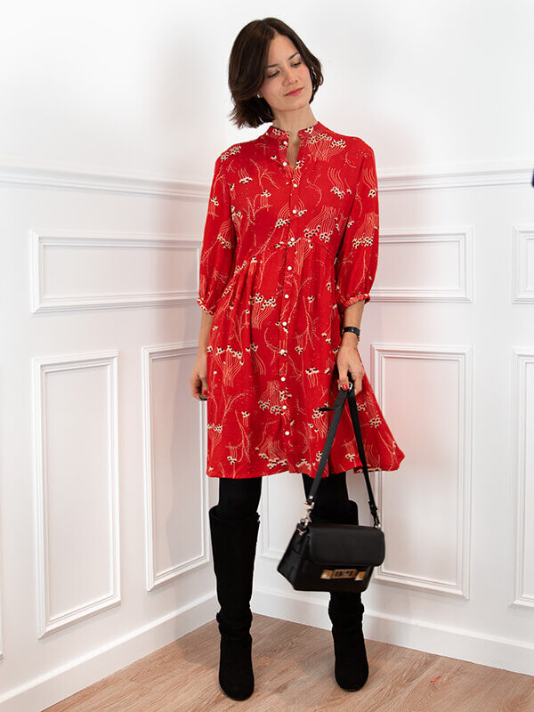 Le patron de robe Pléiades 2 est montré ici dans sa version robe chemise, dans une viscose rouge avec un subtil imprimé floral. La robe est portée par une jeune femme brune dans une pièce bien éclairée avec des moulures haussmanniennes.