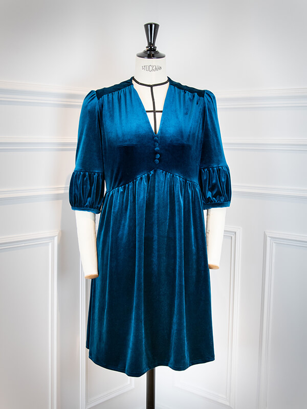 Le patron de robe Pléiades 1 version manches lanternes est réalisée ici en velours bleu. La robe est montrée sur un mannequin blanc, devant un mur blanc à moulures.