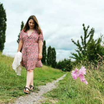 Une jeune femme marche le long d'un chemin dans la nature. Elle a de longs cheveux et porte une robe courte boutonnée devant, légèrement décolletée, à petites manches froncées, réalisée dans un tissu de viscose à imprimé fleuri.