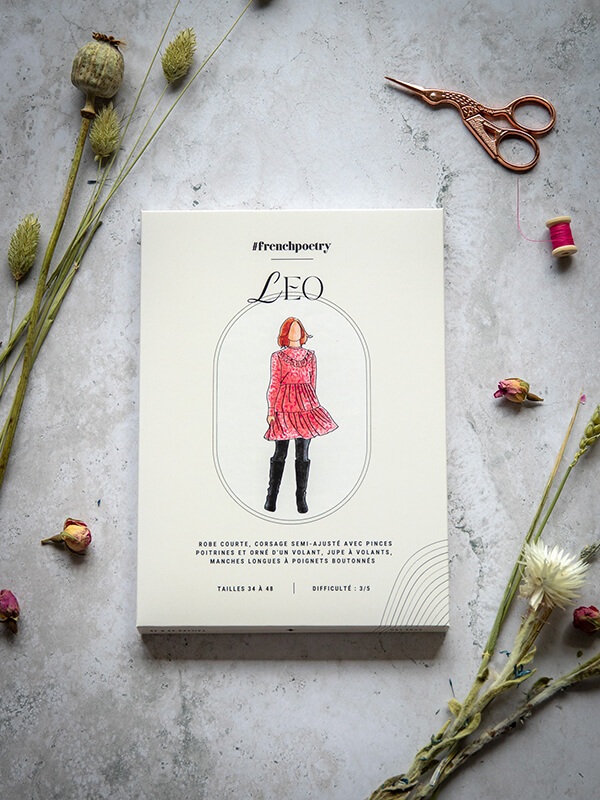 Le patron de robe babydoll Leo est photographié ici au format imprimé A0 avec sa jolie pochette. Il est posé sur un fond marbré parsemé de boutons de rose séchés et de graminées, avec une lumière très douce.