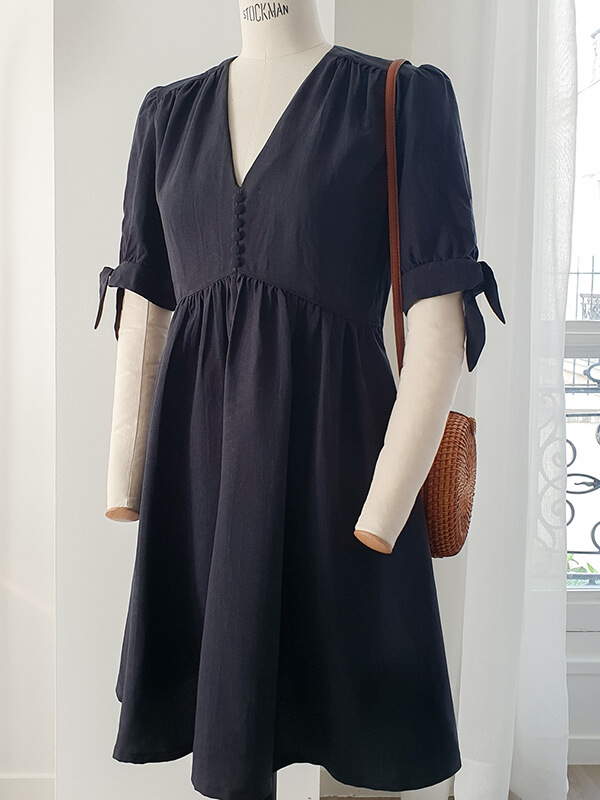 Cette image montre la robe Pléiades 1 réalisée dans un crêpe noir avec des manches courtes nouées sur le côté. Elle est portée par un mannequin de couture, accessoirisée par un petit sac à main en paille.