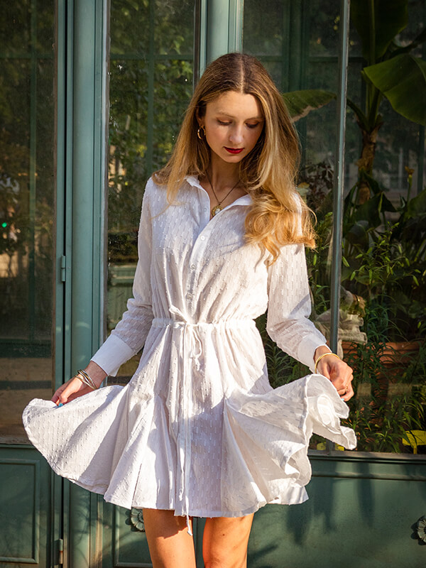 Cette image montre la robe Stella portée par une jeune femme aux longs cheveux blonds ondulés. La robe est réalisée dans un coton blanc très fin, c'est une robe chemise cintrée par une ceinture à coulisse, avec une jupe à godets qui vole au vent.