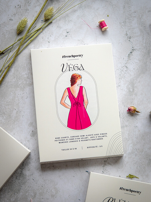 Le patron de robe d'été Vega est photographié ici au format imprimé A0 avec sa jolie pochette. Il est posé sur un fond marbré parsemé de boutons de rose séchés et de graminées, avec une lumière très douce.